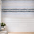 A wall mural or wallpaper featuring a horizontal beach-inspired design with light blue and white textured stripes above a white shiplap lower wall, shown in a styled room with a sofa, pillow, and window.