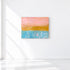Postcard from the Pacific Coast original painting with a pink sky, golden shoreline, and turquoise water, in a white wall.