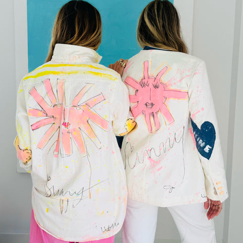 Two models wearing matching hand-painted white jackets with pink sun-face designs and 'Sunny' script lettering. Handpainted by Kerri Rosenthal
