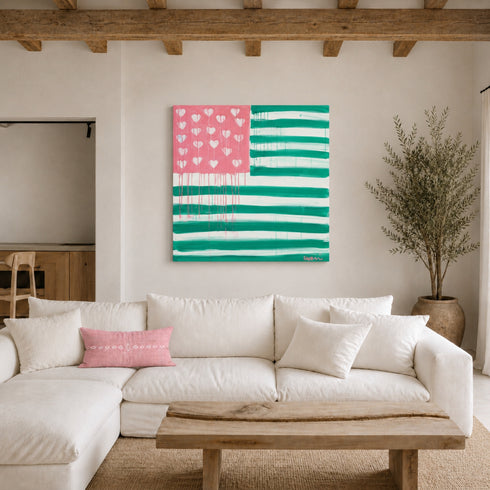 The Heart-Flag painting in pink and green hanging unframed on a plaster wall in a rustic-modern living room with exposed wood beams, above a white linen sectional sofa with a pink embroidered lumbar pillow, a live-edge wood coffee table, and a tall potted olive tree. Framed in plexi, painted by artist Kerri Rosenthal.