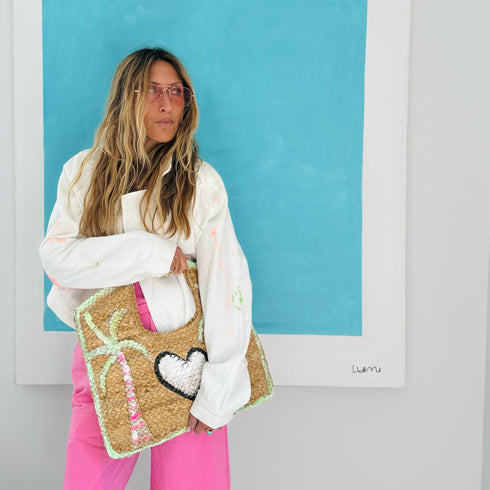 Model wearing a white paint-splattered denim jacket and pink pants, carrying a woven straw tote bag with a palm tree and heart appliqué design. Handpainted by Kerri Rosenthal