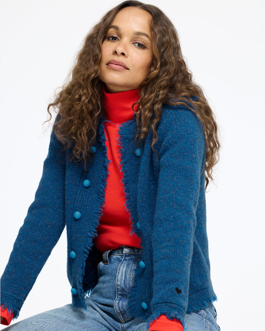 Model seated wearing the Fringe Beau-Lero in electric blue over the Mock Neck Rib Tee in red with jeans, front view