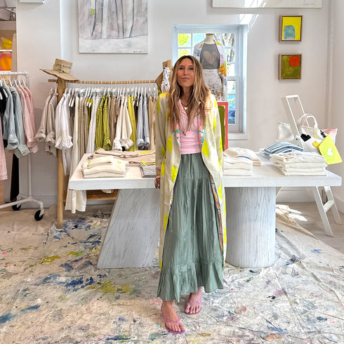 Woman standing in a boutique wearing a cream and neon yellow heart print maxi duster coat with pink floral trim, pink tank top, and sage green tiered maxi skirt.