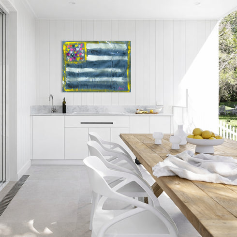 Outdoor kitchen with a wooden dining table and white chairs, featuring a colorful Abstract Flag painting on aluminum with blue, white, and multicolored heart design on a white background. Artwork by Kerri Rosenthal.