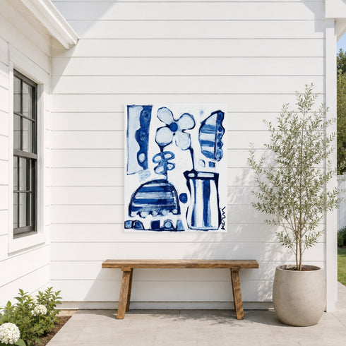 Aluminum outdoor art by Kerri Rosenthal blue and white abstract floral canvas mounted on white shiplap siding above a wooden bench, beside a potted olive tree on an outdoor patio.