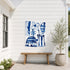 Aluminum outdoor art by Kerri Rosenthal blue and white abstract floral canvas mounted on white shiplap siding above a wooden bench, beside a potted olive tree on an outdoor patio.
