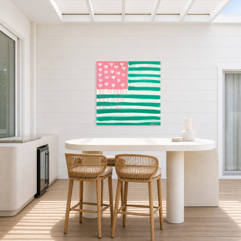 The Heart-Flag aluminum painting in pink and green hanging on a white shiplap wall in an outdoor dining area, above a round white pedestal table with woven rattan counter stools and a built-in beverage cooler nearby. Outdoor art by Kerri Rosenthal.