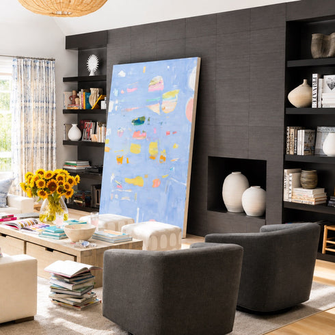 A modern living room featuring a large, colorful abstract canvas painting leaning against a dark gray wood-paneled wall. The room is styled with a cream sectional sofa, two dark gray armchairs, a light wood coffee table, and built-in black shelving.