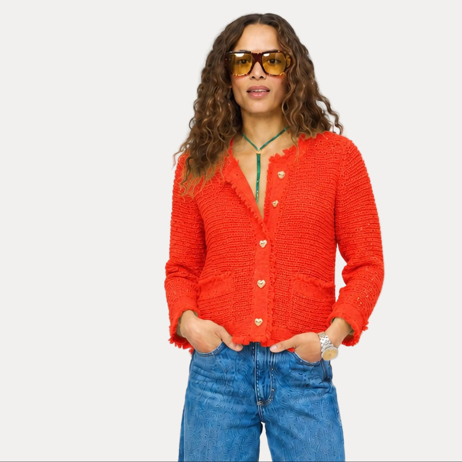A woman modeling a textured orange-red knit cardigan with gold buttons, paired with blue jeans and gold-framed sunglasses, against a plain light gray background.
