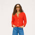 A woman modeling a textured orange-red knit cardigan with gold buttons, paired with blue jeans and gold-framed sunglasses, against a plain light gray background.