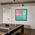 The Heart-Flag painting in pink and green, framed in black, displayed on a wall in a game room. Painted by artist, Kerri Rosenthal.