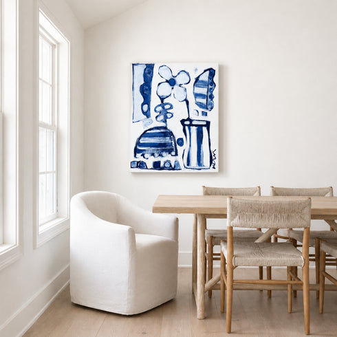Canvas blue and white abstract floral canvas hanging in a dining room above a wood table with woven rush-seat chairs and a white slipcover armchair near a sunlit window.