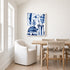 Canvas blue and white abstract floral canvas hanging in a dining room above a wood table with woven rush-seat chairs and a white slipcover armchair near a sunlit window.