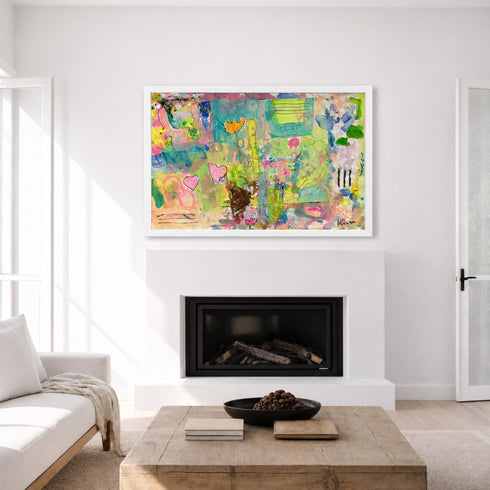 A colorful abstract framed art print hangs on a white wall above a fireplace in a bright, modern living room with a white sofa and wooden coffee table.