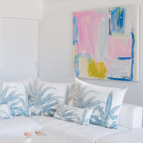 A bright, modern living room featuring a white sectional sofa with blue and white palm-patterned pillows, a round white coffee table, two woven jute poufs on a light rug, and a colorful abstract painting on the white wall.