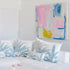 A bright, modern living room featuring a white sectional sofa with blue and white palm-patterned pillows, a round white coffee table, two woven jute poufs on a light rug, and a colorful abstract painting on the white wall.