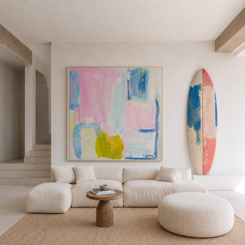 A large abstract canvas painting with pastel pink, blue, and yellow brushstrokes hangs on a white wall above a cream-colored sectional sofa. A surfboard-style art print leans against the wall on the right, and the room is styled with a woven rug, round ottoman, and small wooden side table.