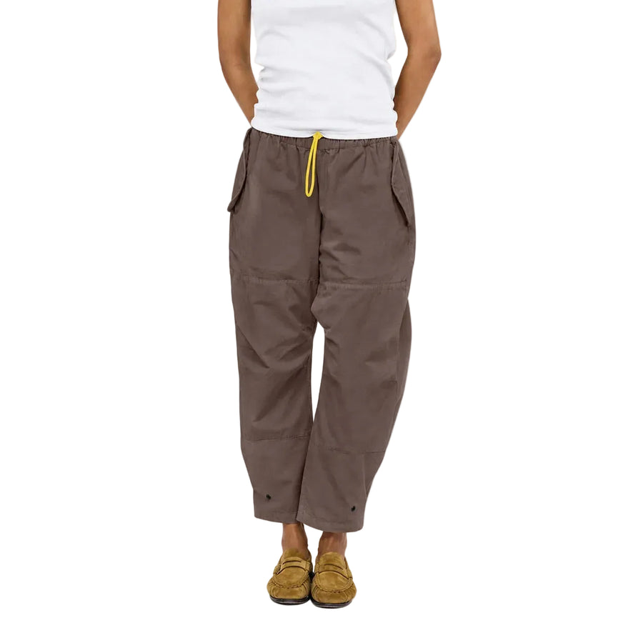Close-up of a model wearing the Love Brigade Pant in brown with a yellow drawcord, a white tank top, and tan shoes, waist-down front view