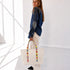 A woman stands in a bright white room holding a cream canvas tote bag with colorful striped handles. She is wearing a blue denim jacket, white pants, and black shoes.