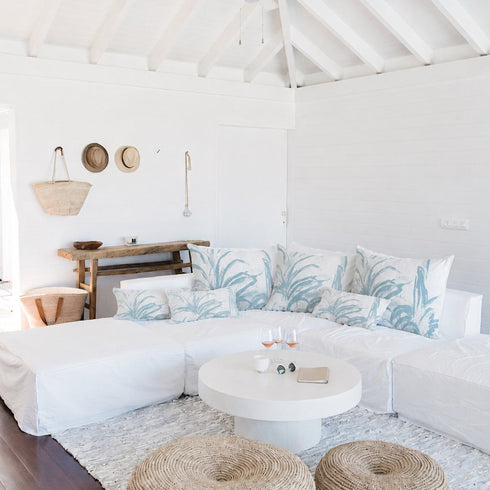 A bright, airy living room with a white sectional sofa decorated with blue and white palm leaf patterned pillows, a round white coffee table, two woven jute poufs on a light rug, and a high white-beamed ceiling.