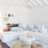 A bright, airy living room with a white sectional sofa decorated with blue and white palm leaf patterned pillows, a round white coffee table, two woven jute poufs on a light rug, and a high white-beamed ceiling.