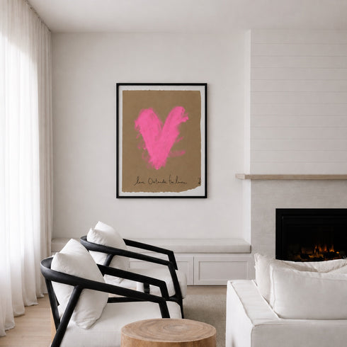 A framed art print featuring a bright pink textured heart on a tan background, hanging on a white wall in a modern living room setting with a fireplace and white furniture.