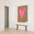 A framed art print featuring a textured pink heart on a brown background, hung on a white wall above a rustic wooden bench in a minimalist room with light wood floors.