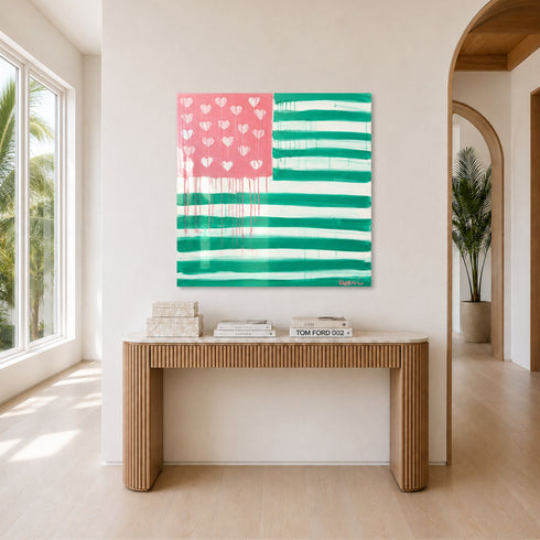 The Heart-Flag painting in pink and green hanging unframed in a bright entryway, above a fluted wood console table styled with stacked design books, near a tall potted palm and arched doorway leading to a sunlit hallway. Framed in plexi, painted by artist Kerri Rosenthal.
