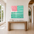 The Heart-Flag painting in pink and green hanging unframed in a bright entryway, above a fluted wood console table styled with stacked design books, near a tall potted palm and arched doorway leading to a sunlit hallway. Framed in plexi, painted by artist Kerri Rosenthal.