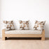 A light wood daybed with a cream upholstered seat cushion and three matching patterned throw pillows, set against a plain white wall on a light wood plank floor.