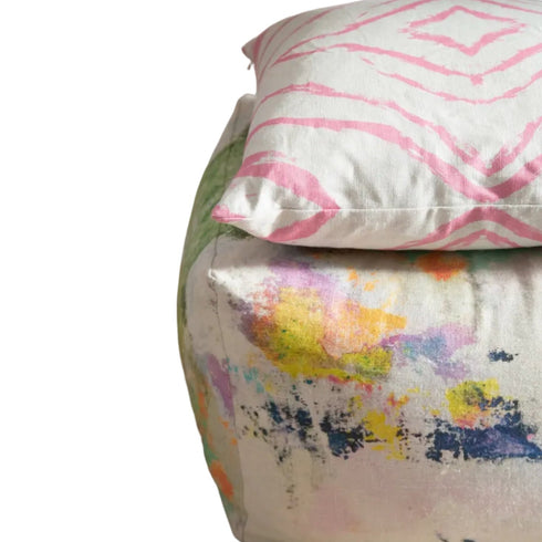 A close-up of a pouf ottoman and matching pillow featuring a colorful abstract watercolor print on an off-white base, with a pink patterned pillow resting on top.