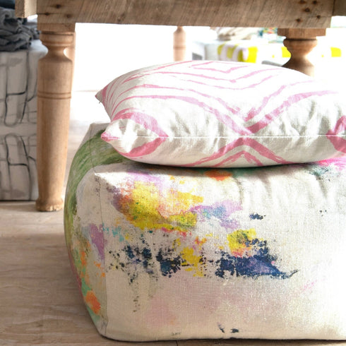 A square upholstered ottoman with a colorful abstract paint-splatter pattern in yellow, blue, green, and pink on a light beige fabric base, with a matching square throw pillow featuring pink wavy lines on a white background resting on top. The ottoman is placed on a light wood floor beneath a wooden table.