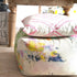 A square upholstered ottoman with a colorful abstract paint-splatter pattern in yellow, blue, green, and pink on a light beige fabric base, with a matching square throw pillow featuring pink wavy lines on a white background resting on top. The ottoman is placed on a light wood floor beneath a wooden table.