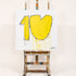 Yellow Kerri Rosenthal abstract painting on canvas of the number '10' next to a drippy heart shape, hand-painted with visible brushstrokes, charcoal outlines, and drip details, signed by the artist in the bottom corner, displayed on a white gallery wall with black dot-patterned accent wall in background. Celebrate ten year anniversary