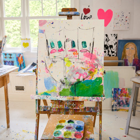 A colorful abstract canvas painting featuring stylized faces and paint splatters, displayed on a white wall in a bright room with white floorboards.