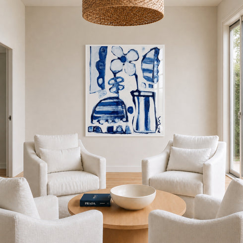 Large framed blue and white abstract floral painting by artist Rosenthal displayed above a round wooden coffee table in a neutral, light-filled living room with white linen armchairs and a woven pendant light.