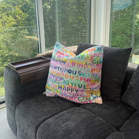 Colorful pillow with text on a gray sofa by a large window with a view of trees.