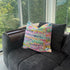 Colorful pillow with text on a gray sofa by a large window with a view of trees.
