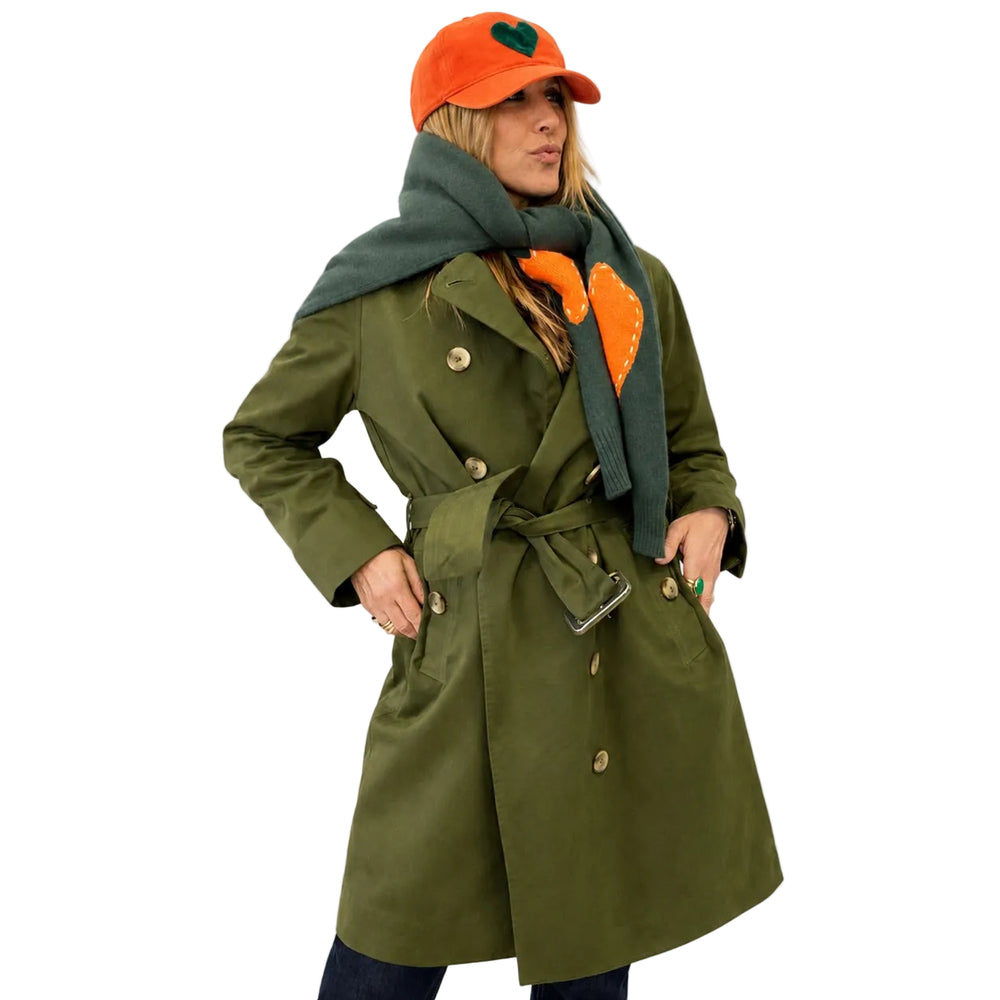 A woman wearing an olive green double-breasted trench coat with a matching belt, a dark green scarf with an orange heart, and an orange baseball cap with a green heart, posed against a plain white background.