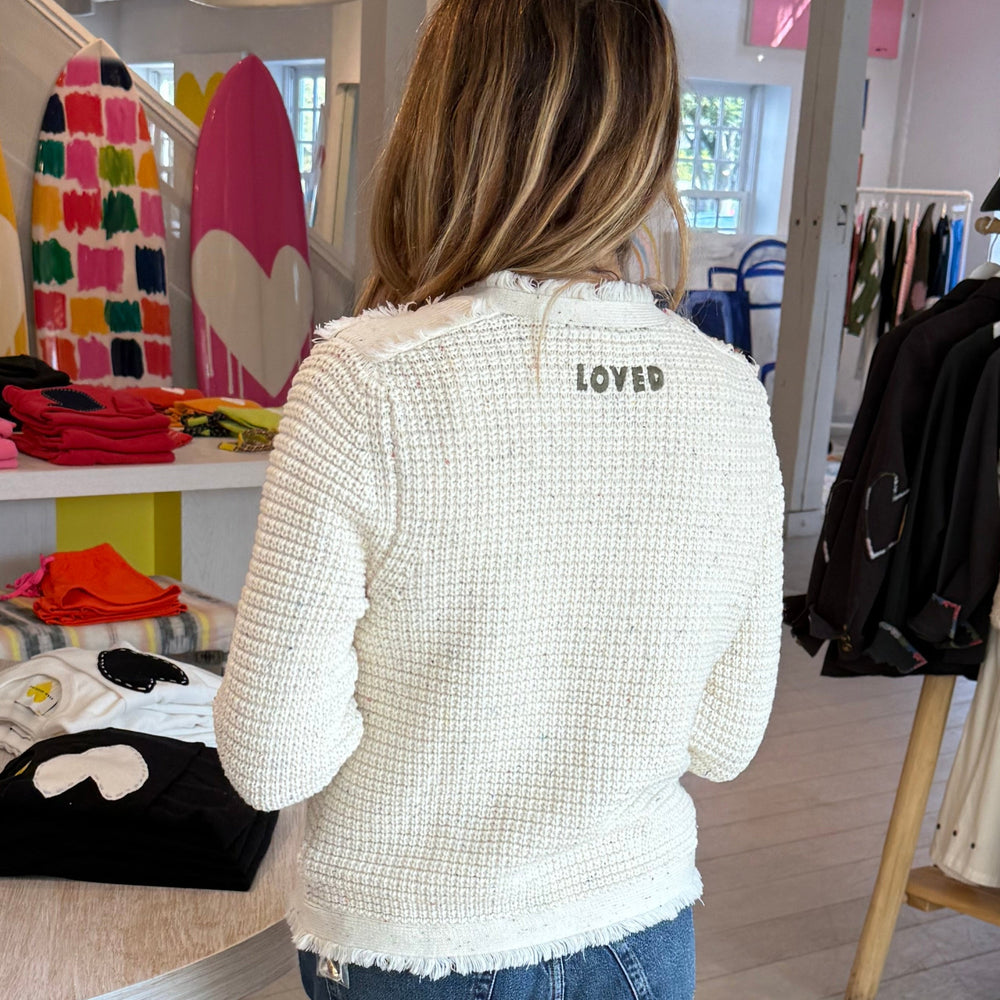 Back of woman wearing a Kerri Rosenthal cream boucle tweed fringe jacket with gold heart buttons, styled with a white tank with pink heart and jeans