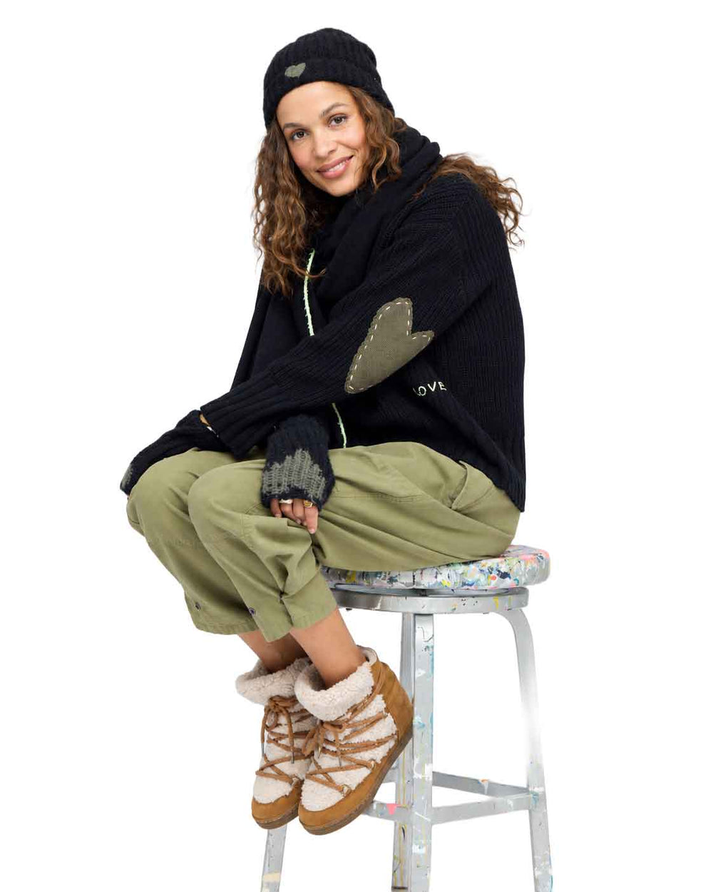 Model sitting on a stool wearing Marlow Imperfect Heart Sweater in black with army green elbow patch, black smittens with grey heart and black winter hat with grey heart detail.