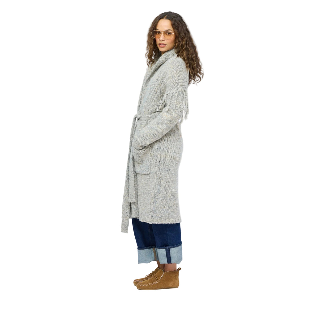 Side profile of model wearing a long gray fringe cardigan belted at the waist, cuffed jeans and tan boots.