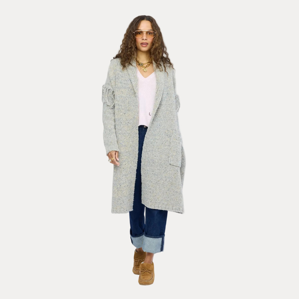 Model walking in a long gray fringe cardigan layered over a white top and cuffed jeans, tan ankle boots, self-tie belt at the waist. Kerri Rosenthal women's sweaters