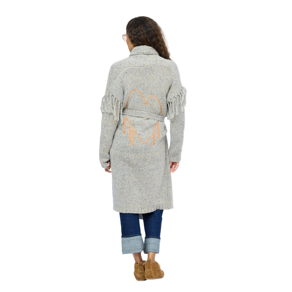 Back view of model wearing a long gray fringe cardigan with an orange open heart embroidered between the shoulder blades, cuffed jeans and tan boots.