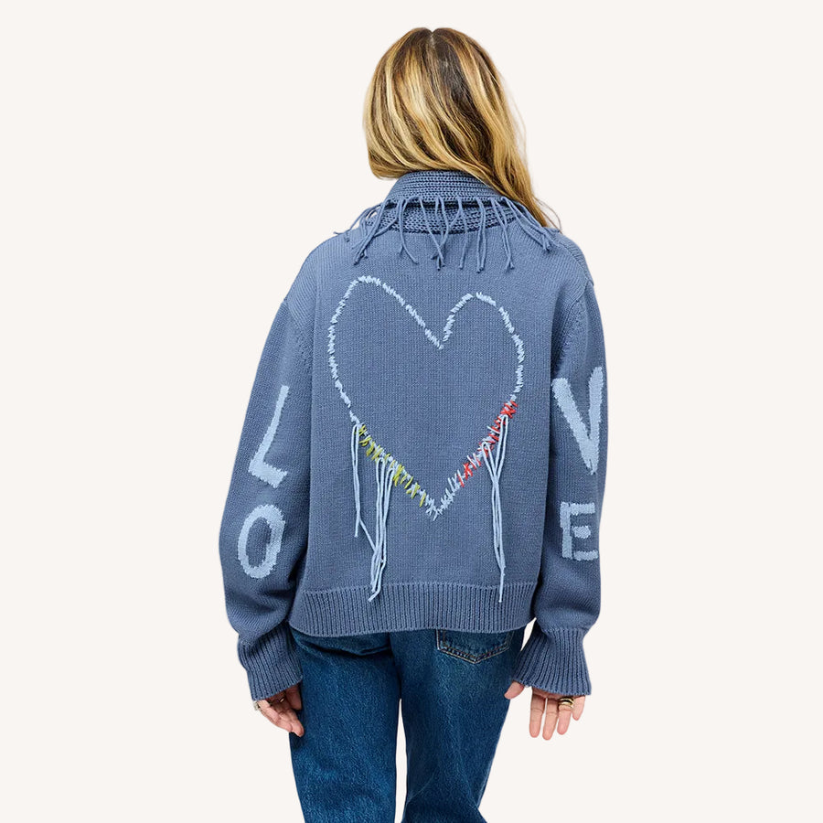 Back view of a woman wearing a Kerri Rosenthal slate blue knit cardigan featuring a large intarsia heart, colorful fringe detail, and 'LOVE' lettering on the sleeves, styled with blue jeans.