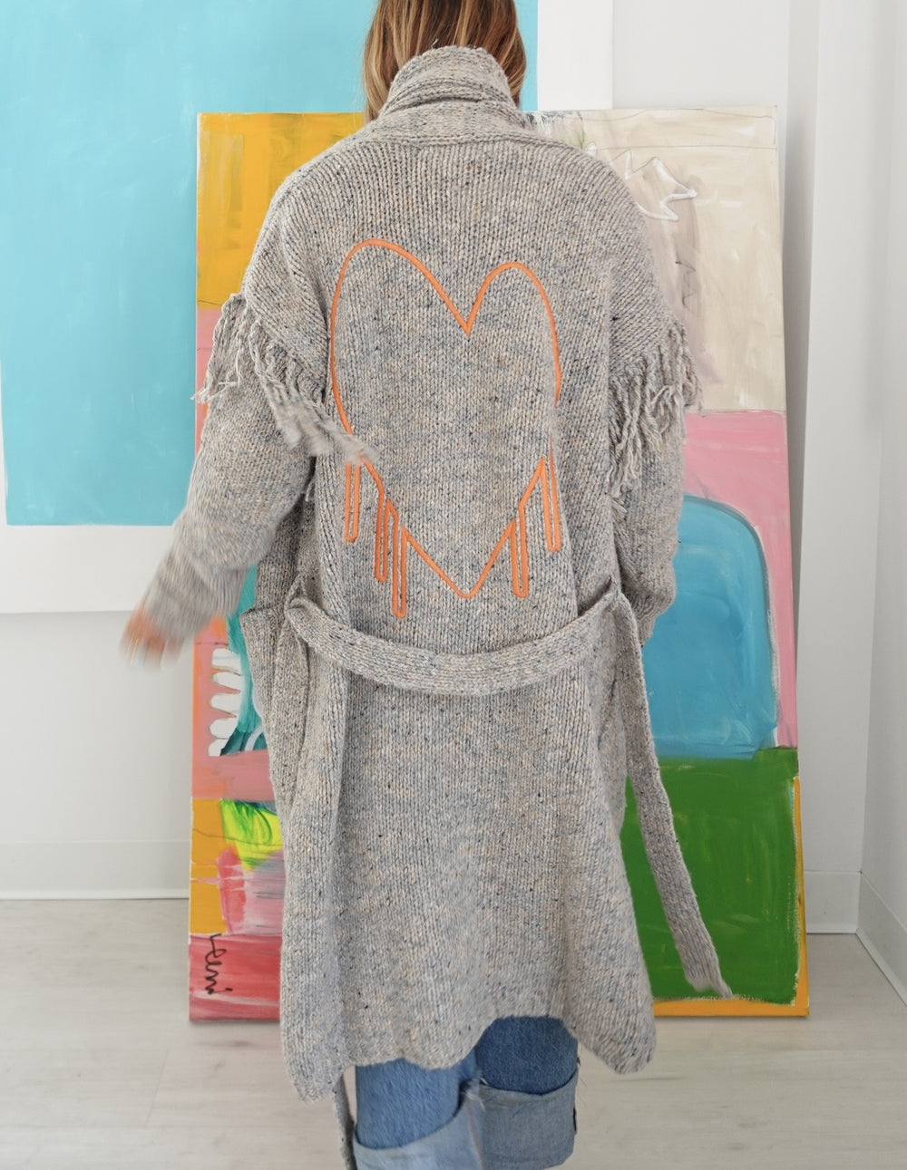 Back view of model wearing a long gray fringe cardigan with an orange open heart embroidered between the shoulder blades,