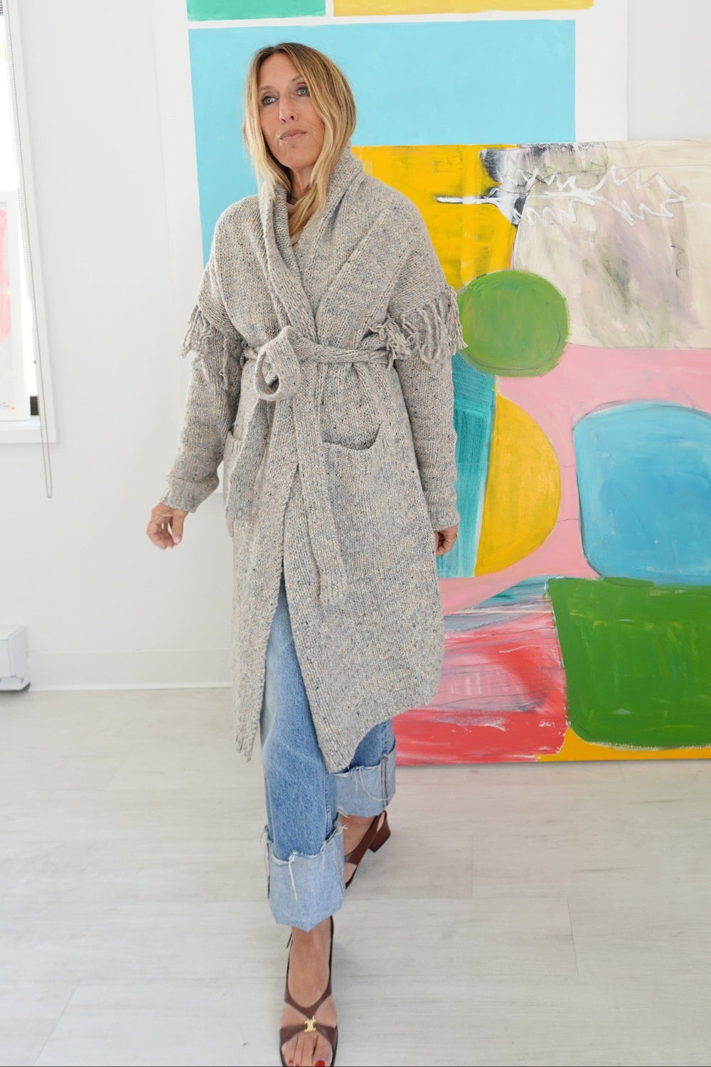 Model wearing a long gray fringe cardigan belted at the waist, cuffed jeans.