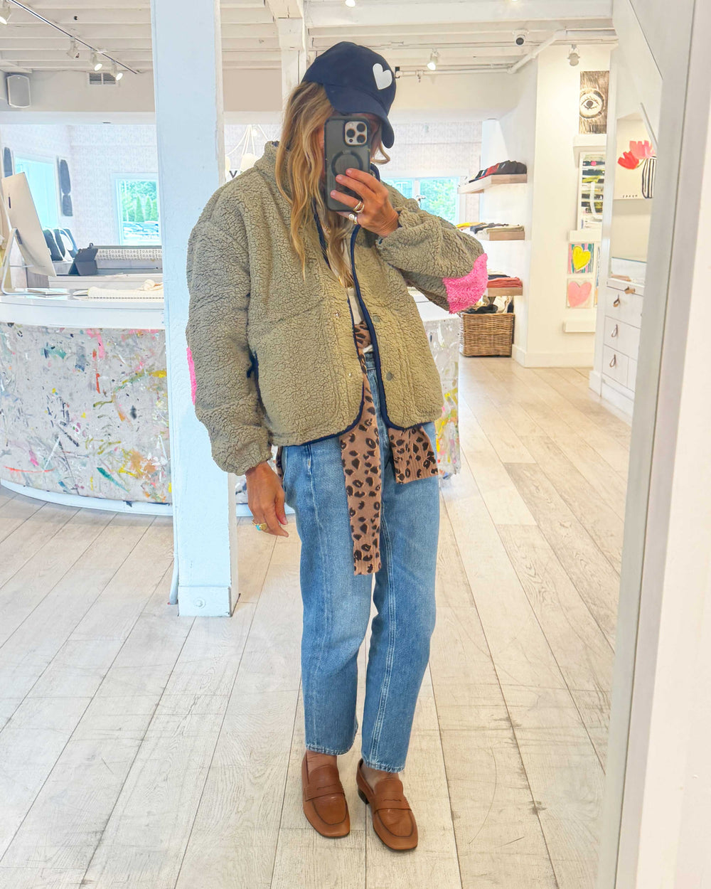 Kerri in an olive teddy fleece jacket with pink color-block trim, jeans, and a navy heart baseball cap, mirror selfie in the Kerri Rosenthal store.