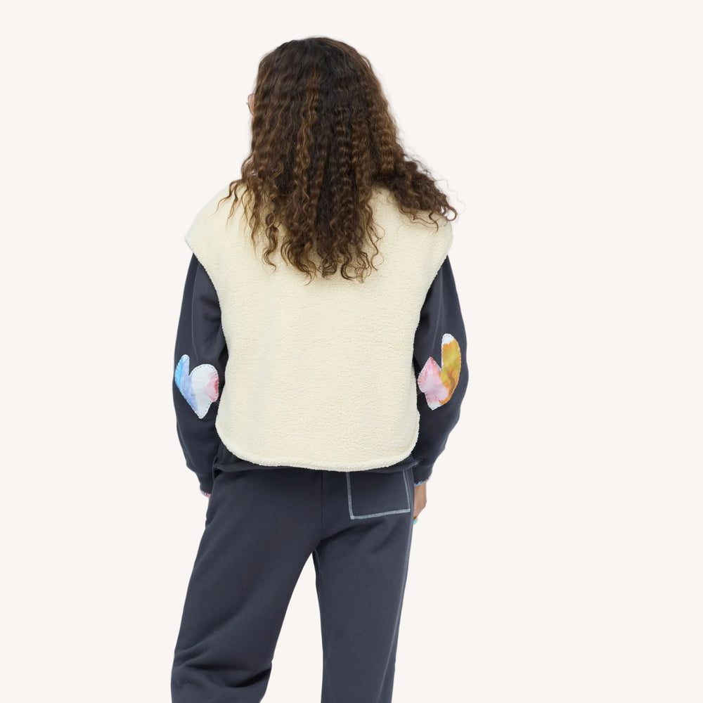 Back view of woman wearing a cream sherpa vest with colorful patchwork sleeves over a navy sweatshirt and navy sweatpant.