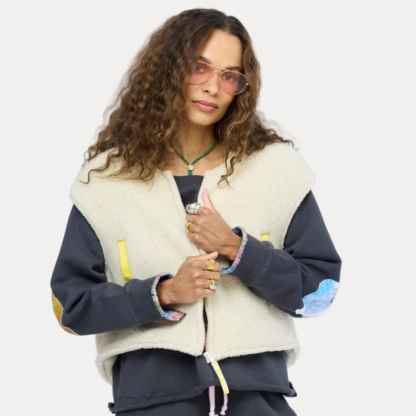 Woman wearing a cream sherpa vest with colorful patchwork sleeves over a navy studio sweatshirt, with sunglasses and a necklace.